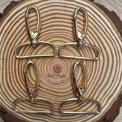 Quatro mosquetões dourados sobre superfície de madeira com selo Alice Prado