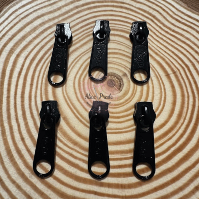 Seis puxadores de zíper pretos sobre superfície com padrão de círculos concêntricos em tons de bege e castanho