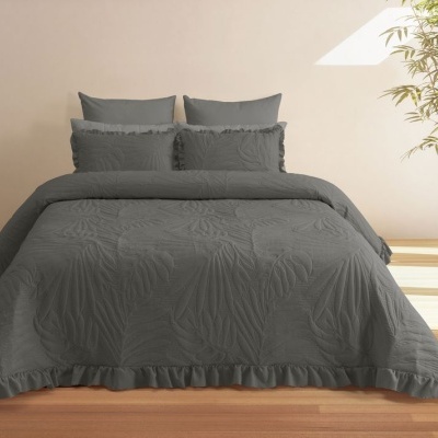 Conjunto de cama cinzento escuro com edredão e almofadas com textura e folhos, numa sala com chão de madeira e planta em vaso branco.