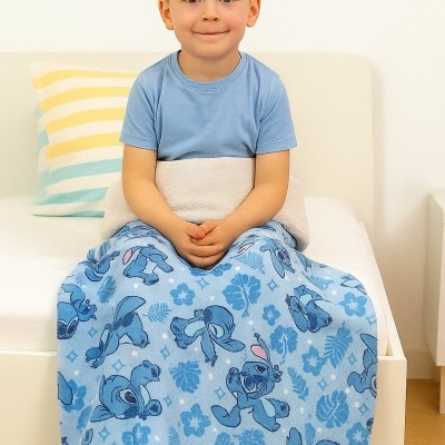 Menino sentado na cama com manta azul estampada de Stitch e almofada às riscas.