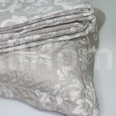 Cobertor bege texturizado com padrão floral branco dobrado sobre mesa branca ao lado de vaso de plantas