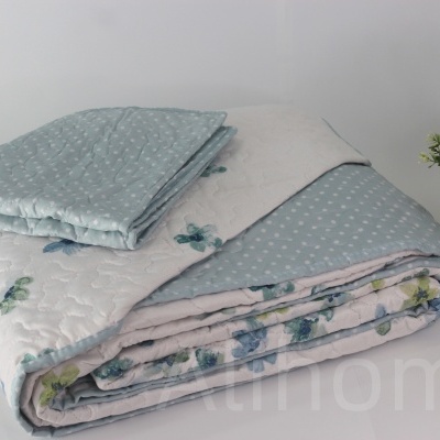 Colcha acolchoada estampada azul e branca com flores e estrelas enrolada ao lado de vaso dourado com planta