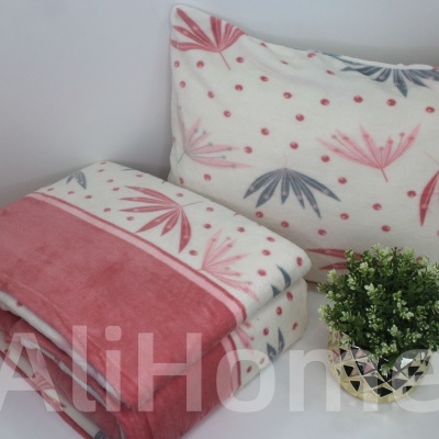 Almofada e manta decorativa com padrão de folhas rosa e cinza ao lado de vaso com planta verde.