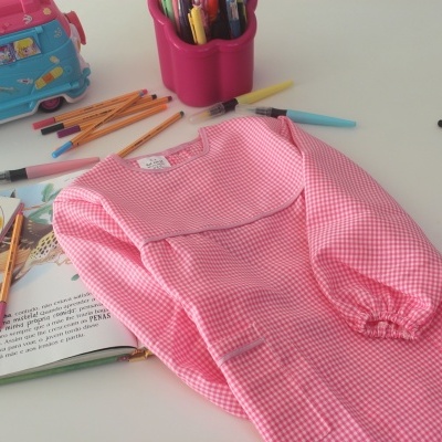 Avental rosa xadrez para criança com livro aberto, lápis de cor e estojo rosa na mesa branca