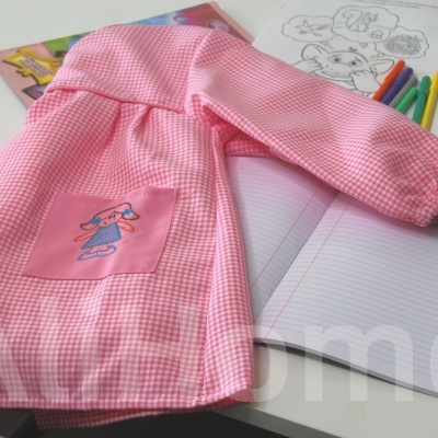 Avental de criança rosa com padrão vichy e bolso com desenho de menina sobre mesa com materiais de desenho