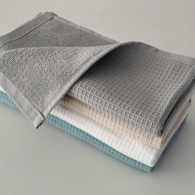 Conjunto de quatro toalhas empilhadas em cores cinza, bege, branco e azul, com textura waffle.