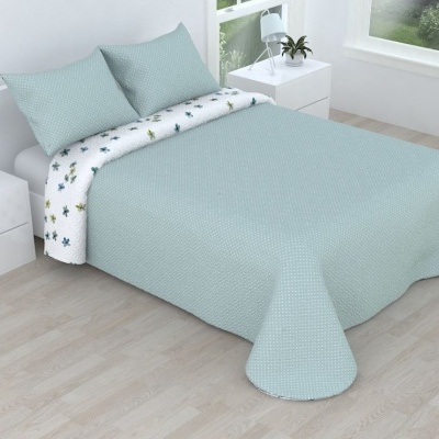 Cama de casal com colcha azul clara matelassada e lençol com padrão de folhas verdes