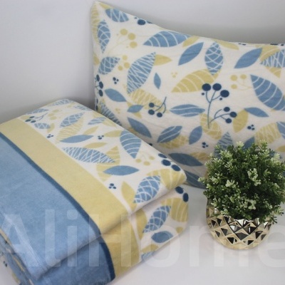 Conjunto almofada e manta com padrão de folhas azul e amarelo junto a vaso com planta artificial.