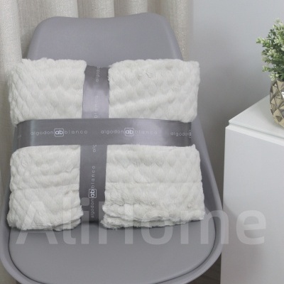 Manta branca macia com padrão de tranças numa cadeira cinzenta, com embalagem de fita cinzenta com texto.