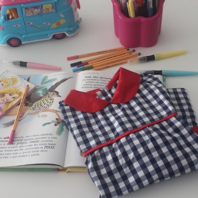 Vestido infantil xadrez azul e branco com gola vermelha sobre livro aberto e lápis de cor