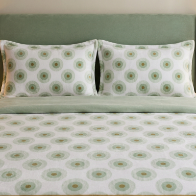 Cama com cabeceira verde água, roupa de cama branca com padrão circular verde e bege, duas mesas de cabeceira em madeira com lâmpadas