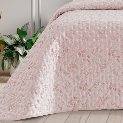 Colcha rosa clara com padrão floral em relevo numa cama ao lado de plantas e mesa de madeira