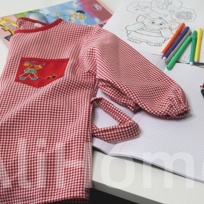 Avental infantil vermelho e branco com bolso vermelho sobre mesa com material de desenho