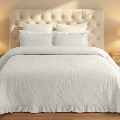 Conjunto de cama branco com padrão floral e folhos, cabeceira bege estofada, mesas de cabeceira de madeira clara e lâmpadas em quarto iluminado
