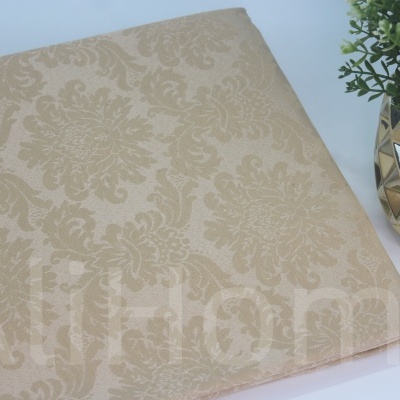 Toalha bege clara com padrão floral e vaso dourado com planta artificial ao lado