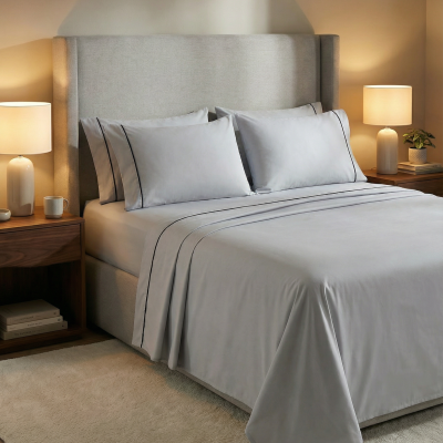 Cama com cabeceira cinza, jogo de cama cinza com linhas pretas, mesas de madeira com lâmpadas, livros e planta, tapete claro