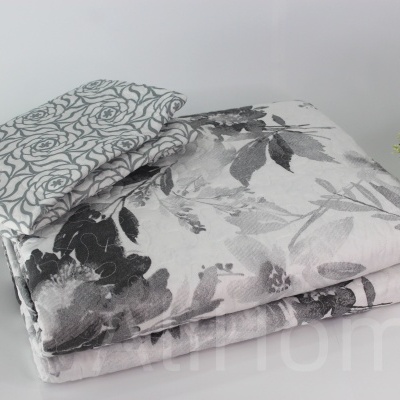 Conjunto de roupa de cama cinzenta com padrão floral preto e branco ao lado de vaso dourado com planta
