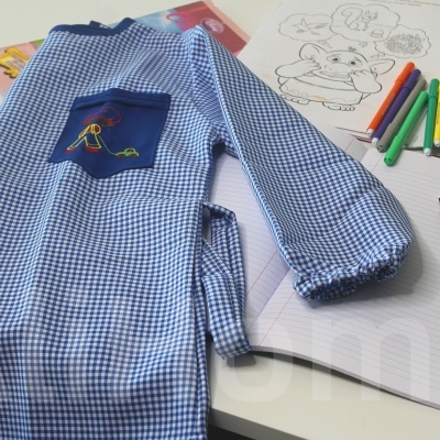 Avental infantil azul e branco às quadrículas com bolso azul e desenhos, sobre mesa com livros de colorir e caderno