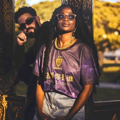 Camisola preta com padrão galáctico e texto em chinês e português, mulher com calças de ganga e óculos de sol.