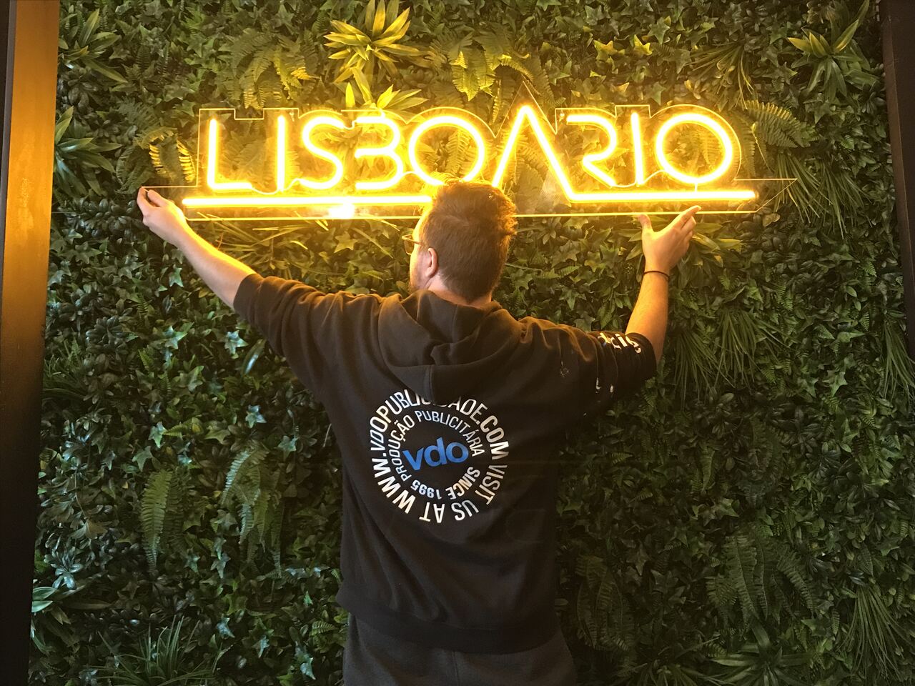 Letreiro de néon amarelo LISBOARIO numa parede verde com pessoa de costas vestindo camisola preta.