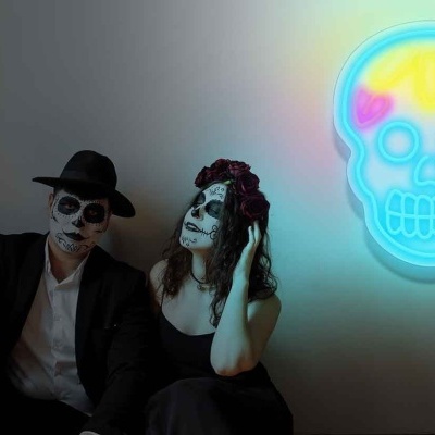 Duas pessoas com maquiagem de caveira sentadas junto a uma parede com luz neon colorida em forma de caveira.