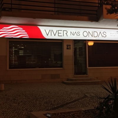 Fachada de loja com letreiro 'VIVER NAS ONDAS' iluminado à noite.