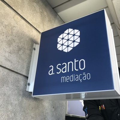 Sinal exterior azul com logotipo branco e texto 'a. santo mediação' fixado numa parede cinzenta