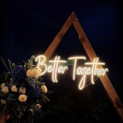 Letreiro luminoso 'Better Together' numa estrutura de madeira triangular com flores e ramos verdes
