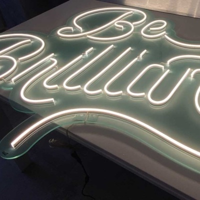 Letreiro luminoso branco 'Be Brilliant' em suporte transparente sobre superfície clara
