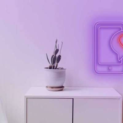 Luz de néon em forma de telemóvel com coração numa parede branca junto a uma cadeira branca e um armário com vaso e planta.