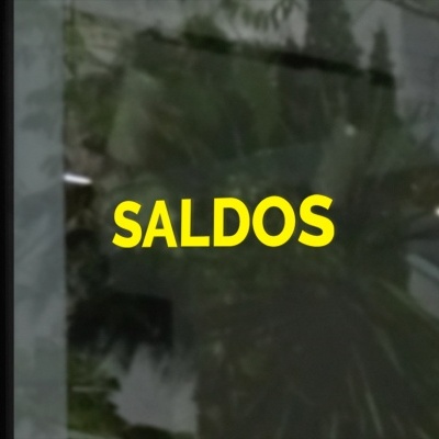 Letreiro amarelo com a palavra SALDOS num vidro com reflexos de planta e escada