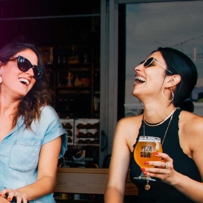 Duas mulheres a rir a beber cerveja junto a letreiro neón cor-de-rosa