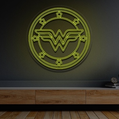 Símbolo néon amarelo com logótipo da Mulher Maravilha numa parede cinzenta, móvel de madeira e plantas em vasos