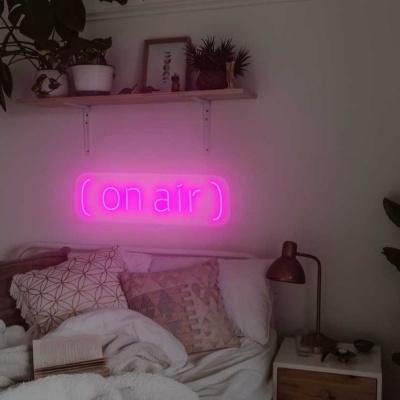 Quarto com cama branca, almofadas neutras, plantas, candeeiros de cabeceira e placa de néon rosa '(on air)'}