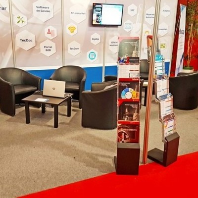 Espaço de exposição com cadeiras pretas, mesas pretas com portátil aberto e expositores de folhetos em fundo azul e branco.