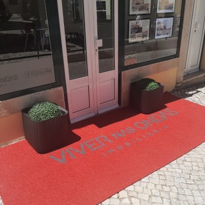 Tapete vermelho com texto cinza à entrada de loja imobiliária com flores artificiais e portas brancas