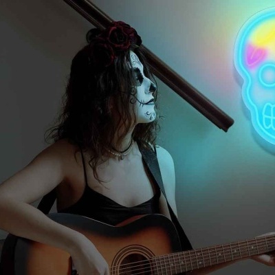 Mulher maquilhada de caveira tocando guitarra acústica junto a mesa com velas e néon de caveira colorido na parede