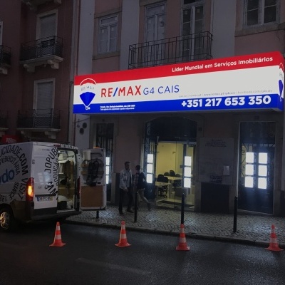 Agência imobiliária RE/MAX G4 CAIS com letreiro iluminado e veículos estacionados à noite