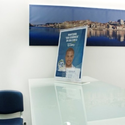 Suporte de mesa transparente com imagem publicitária e texto em português sobre mesa branca brilhante com cadeiras azuis e fotografia panorâmica na parede