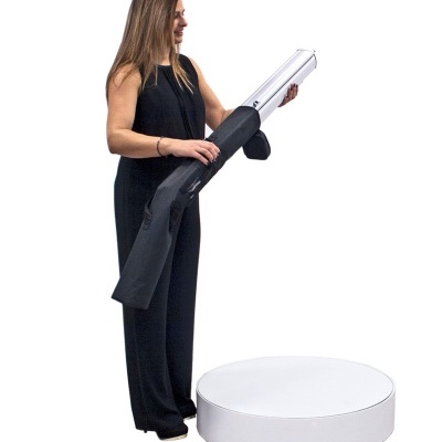 Mulher com macacão preto segurando capa preta com objeto cilíndrico prateado, em fundo branco.