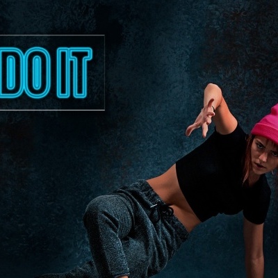 Pessoa em pose dinâmica com gorro rosa, camisa preta e calças de ganga, fundo escuro com texto JUST DO IT em azul