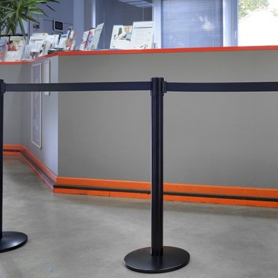 Sistema de barreiras de filas com postes e fitas pretas em espaço interior com balcão cinza e linha laranja