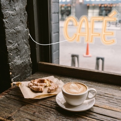 Chávena de café com leite e pastel de nata sobre mesa de madeira junto à janela com letreiro 