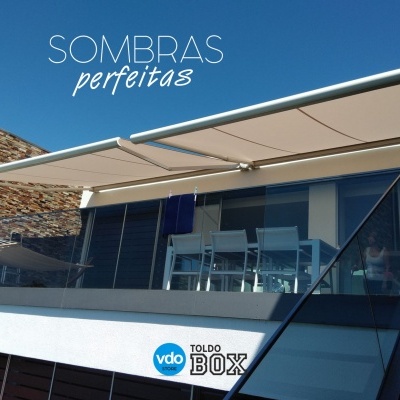 Toldo retrátil bege numa varanda moderna com mesa e cadeiras brancas, texto SOMBRAS perfeitas, vdo TOLDO BOX