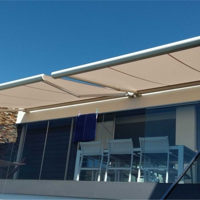 varanda com toldo retrátil bege, mesa e cadeiras azuis, guarda-corpo de vidro e parede de pedra