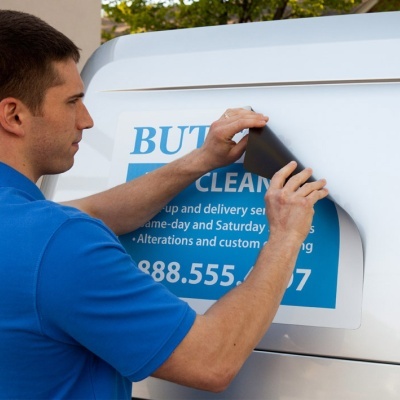 Homem aplicando autocolante azul e branco numa carrinha branca com texto e número visível