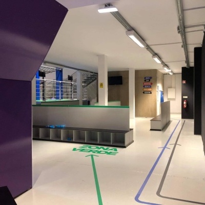 Espaço interior com chão branco, linhas coloridas, bancadas e sinais no fundo.