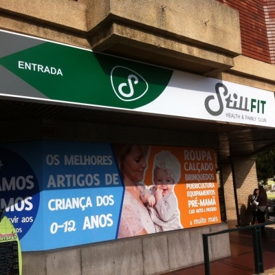 Fachada com entrada Still Fit Health & Fitness Club, letreiro verde e branco, cartaz colorido com texto em português sobre artigos para crianças
