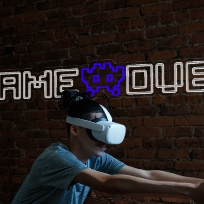 Criança usa headset VR branco na frente de parede de tijolos com texto GAME OVER