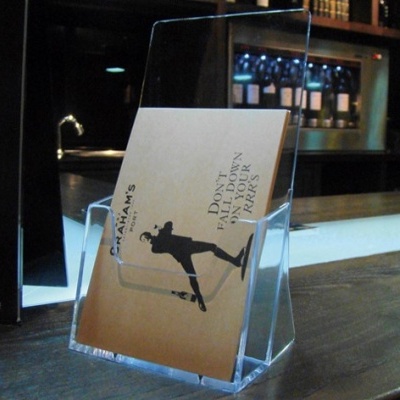 Expositor acrílico com folheto publicitário numa superfície de bar em madeira.